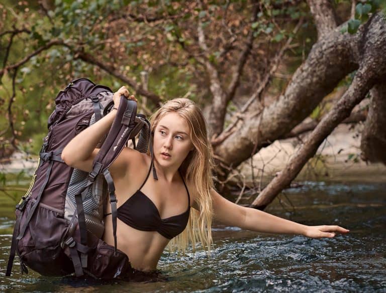 A young woman holding her backpack high when crossing a mountain creek in the Sierra Nevada Mountains of Northern California.