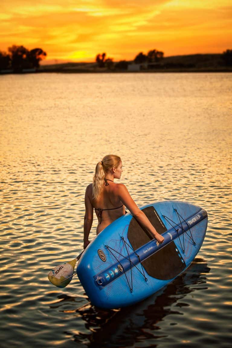 A young woman wading into a small lake with a paddle board at sunset.