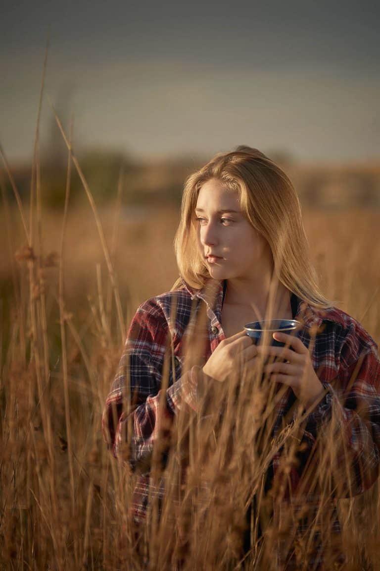 A young woman in a flannel shirt drinking coffee outdoors in a field.