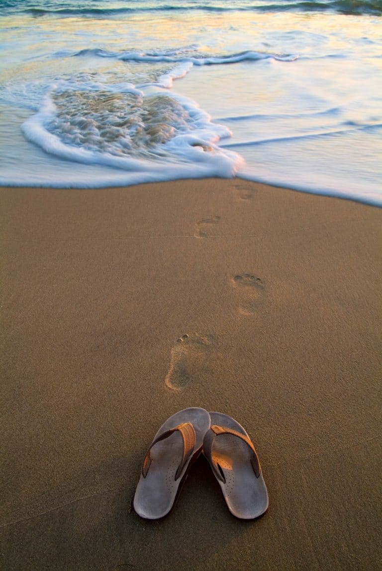 Sandals on a beach in Southern California with footprints in the sand leading towards the water.