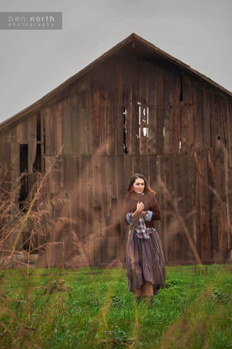 A young woman wearing Free People clothing standing in a field next to a barn on a cold, Winter day.