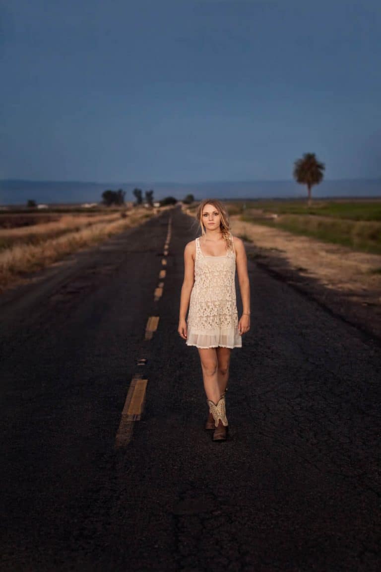 Young woman in cowboy boots and a Free People slip walking down a back road in a rural area on a Summer evening.